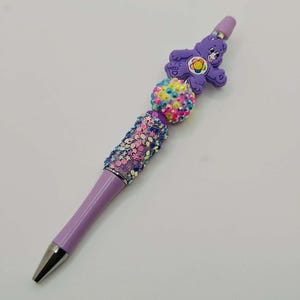 Puede incluir: Un bolígrafo morado con punta plateada, adornado con una figura de Care Bear y detalles de cuentas coloridas. El bolígrafo presenta una sección brillante y una parte superior decorativa, ideal para escribir o coleccionar.