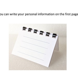 May include: A small, white, lined notepad with black binding. The notepad is standing upright, with the first page visible. The text "You can write your personal information on the first page" is above the notepad.