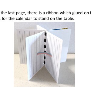May include: A white calendar with lined pages, held open to display its structure. The text "On the last page, there is a ribbon which glued on it. It is for the calendar to stand on the table." is visible. A ribbon is glued to the last page.