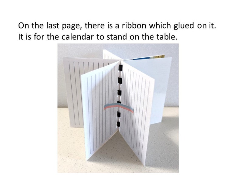May include: A white calendar with lined pages, held open to display its structure. The text "On the last page, there is a ribbon which glued on it. It is for the calendar to stand on the table." is visible. A ribbon is glued to the last page.