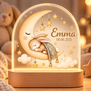 Pode incluir: Uma luz noturna de acrílico em arco com base de madeira apresenta um coelho a dormir numa lua crescente, enrolado numa manta cinzenta. O design inclui estrelas amarelas, nuvens brancas e o texto Emma 08.06.2025.
