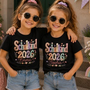 Könnte beinhalten: Zwei junge Mädchen tragen schwarze T-Shirts mit dem Text "Schulkind 2026" in Pastellfarben, zusammen mit den Namen "Emma" und "Sophia". Sie tragen Sonnenbrillen und Jeansshorts. Die Shirts haben Blumen- und Herzakzente.