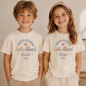Könnte beinhalten: Zwei Kinder tragen cremefarbene T-Shirts mit dem Text "ERSTE KLASSE Schulkind" in einem Regenbogenbogen, einem rosa Herz und den Namen "Karl" und "Lina" darunter. Das Jahr "2026" ist darunter gedruckt. Kurzärmlig.