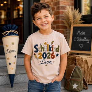 Könnte beinhalten: Ein lächelnder junger Mensch trägt ein beiges T-Shirt mit der Aufschrift "Schulkind 2026 Leon" in bunten Buchstaben. Eine Schultüte und ein Rucksack sind zu sehen. Die Szene deutet auf den ersten Schultag hin.