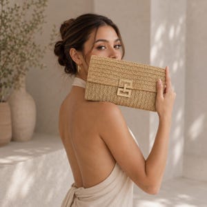 Könnte beinhalten: Eine rechteckige Clutch aus gewebtem Stroh mit einem goldfarbenen Verschluss. Die Clutch hat eine hellbraune Farbe und ein strukturiertes Aussehen. Der Verschluss hat ein geometrisches Design. Die Tasche wird vor einer Person gehalten.