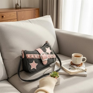 Può includere: Borsa a tracolla in denim nero con dettagli a stella rosa e cinturino in pelle rosa. La borsa presenta una tracolla a catena argentata e una grande chiusura con fibbia. La borsa è su un divano grigio.