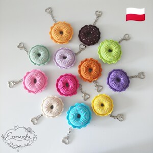 Puede incluir: Una colección de llaveros de donuts de ganchillo de colores, incluyendo rosa, naranja, amarillo, verde y azul. Cada llavero tiene un cierre plateado en forma de corazón. Los llaveros están dispuestos sobre una superficie blanca.