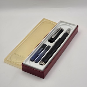 May include: A black Sheaffer fountain pen with a matching cap, two blue ink cartridges, and a translucent plastic case with the Sheaffer brand name embossed on the lid. The pen set is presented in a rectangular box with a maroon base and white interior.