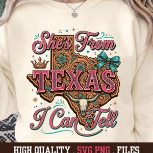 May include: Cream-colored sweatshirt with a graphic design featuring the state of Texas. The design includes the text "She's From Texas I Can Tell" in pink and brown, with floral and steer skull accents. The bottom of the image reads "HIGH QUALITY . SVG PNG . FILES".