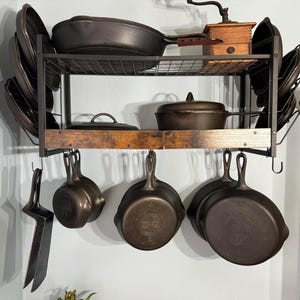 May include: A black metal pot rack with cast iron cookware. The rack holds skillets, pans, and a lid. A wooden spice grinder sits on the top shelf. The cookware is hanging and stacked, showcasing a rustic kitchen aesthetic.