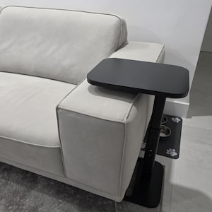 May include: A black, adjustable side table with a rectangular tabletop is positioned next to a light gray sofa. The table has a vertical support and a base. The sofa has a light gray color and a modern design.