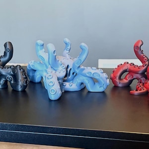 May include: Three octopus figurines arranged in a row, each featuring multiple curved tentacles with visible suction cups. The figurines are coloured in matte black, light blue, and a gradient of dark red to black, all displayed in a standing position.