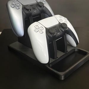 Op de afbeelding: Een zwarte en grijze gamecontrollerstandaard met twee controllers, één zilverkleurig en één wit. De standaard heeft een bakje voor accessoires. De controllers hebben zwarte knoppen en directionele pads. De standaard staat op een zwart oppervlak.