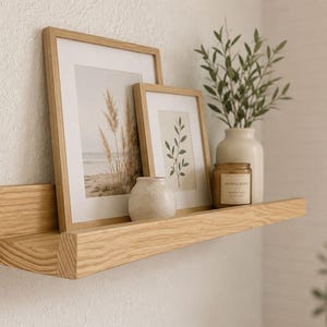 May include: A light-toned wooden floating shelf mounted on a wall, holding two framed prints of botanical and coastal scenes, a small speckled ceramic vase, a glass candle jar with a label, and a white vase containing green leafy stems.