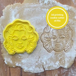 Puede incluir: Un cortador de galletas de plástico amarillo con forma de rostro de mujer, junto a una galleta cortada con la misma forma. El texto "3d printing cookie cutter stl file" está en un círculo amarillo. La galleta y el cortador están sobre una superficie espolvoreada con harina.