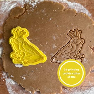 May include: A yellow plastic frog-shaped cookie cutter and a cookie cut-out of the same shape sit on a brown surface. The cookie cutter has a crown. A yellow circle with the text "3d printing cookie cutter stl file" is in the image.