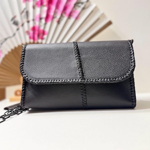 Puede incluir: Un bolso de mano rectangular negro con superficie texturizada y cierre de solapa. Los bordes y la costura central están detallados con costuras negras gruesas y trenzadas. Una fina correa de cadena de metal negro está unida al lateral.
