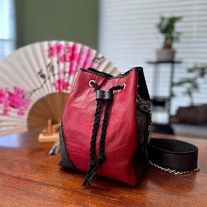 Puede incluir: Un bolso tipo saco confeccionado en cuero rojo y negro, con cierre de cordón ajustable mediante trenzas negras, ojales de metal plateado y una correa de hombro de cuero negro con un eslabón de cadena metálica plateada.