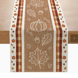 May include: A rectangular table runner featuring a tan burlap-textured centre panel with white line art of pumpkins, maple leaves, and acorns. The side borders display a brown, orange, and cream plaid pattern with small pumpkin and acorn icons.