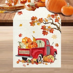 Può includere: Un runner da tavola rettangolare con un'illustrazione stampata di un vecchio pick-up rosso che trasporta varie zucche. Il design include un ramo d'albero con foglie autunnali arancioni e gialle, e la parola WELCOME è scritta sulla sponda posteriore del veicolo.