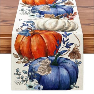 May include: A rectangular table runner featuring a repeating pattern of orange, white, and blue pumpkins. The design includes blue and brown foliage, small white flowers, and thin vines printed on a light beige, textured fabric background.