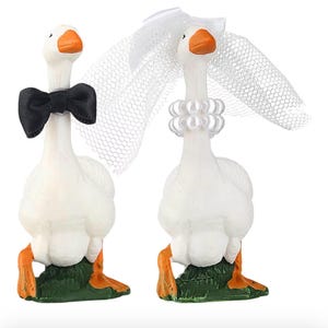 May include: Two white goose figurines, one with a black bow tie and the other with a pearl necklace and a white veil. Both have orange beaks and feet, standing on green bases. Wedding or anniversary themed.
