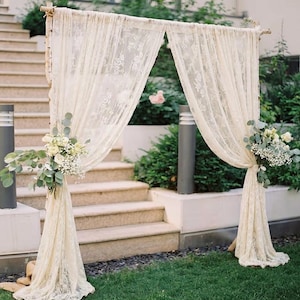 Può includere: Un arco nuziale in pizzo color crema decorato con fiori bianchi e verde. L'arco è drappeggiato con tende di pizzo fluenti e si trova su un prato verde, con gradini di pietra sullo sfondo.