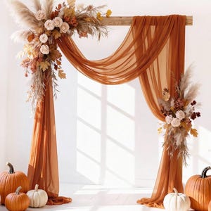 May include: A wooden wedding arch draped with sheer burnt orange fabric, adorned with arrangements of pampas grass, dried foliage, and pale pink roses. Several orange and white pumpkins are positioned on the floor at the base of the arch.