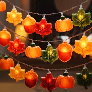 May include: A string of decorative LED lights featuring translucent plastic shapes in the form of orange pumpkins, brown acorns, and maple leaves in shades of yellow, orange, red, and green. Each individual ornament glows with a warm light.