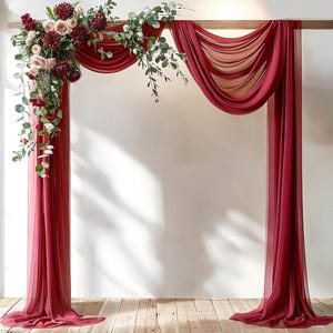 May include: A wedding arch decoration featuring deep red sheer fabric drapes hanging from a horizontal wooden beam. A large floral arrangement of dark red dahlias, light pink roses, and green eucalyptus leaves is attached to the upper left corner.