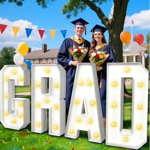 Op de afbeelding: Een afstudeerscène met twee afgestudeerden in marineblauwe toga's en hoeden, met boeketten. Grote witte letters met de tekst "GRAD" met verlichte lampen staan ​​op de voorgrond. Ballonnen en kleurrijke vlaggetjes versieren de achtergrond.