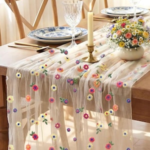 May include: A sheer, cream-colored table runner adorned with colorful embroidered flowers. The runner is draped over a wooden table set with floral-patterned plates, gold cutlery, and a vase of flowers. A lit candle sits in a gold candlestick.