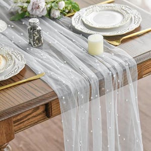 Peut inclure: Chemin de table blanc transparent orné de fausses perles. Il est posé sur une table en bois avec des assiettes blanches, des couverts dorés et une petite bougie. Un arrangement floral est visible en arrière-plan.