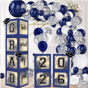 Puede incluir: Un conjunto de decoración para fiesta de graduación con cajas azul marino que deletrean "GRAD" y "2026", adornadas con gorros de graduación. Las cajas están rodeadas de globos azul marino, plateados y blancos, creando un ambiente festivo.