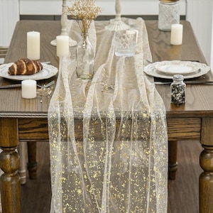May include: A dining table set for a meal, featuring a sheer, gold-flecked table runner. The table is set with white plates, candles, and a croissant. A small glass bottle holds dried flowers, and a glass of water is visible.