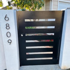 May include: A black metal gate with horizontal rectangular cutouts. The gate is part of a fence with the address numbers 6809 displayed on the left side. The gate is set in a concrete and brick walkway, with greenery visible through the cutouts.