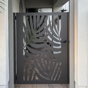 May include: A dark gray metal gate with a leaf design cut out. The gate has a black handle and is set in a doorway. The gate is made of metal and is a decorative element for a home's entrance.