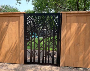 May include: A black metal gate with a tree branch design is set between two wooden fence panels. The gate features a handle and a latch. The background shows a green lawn and trees.