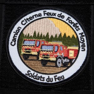 Puede incluir: Parche circular con el texto "Camion Citerne Feux de Forêts Moyen Soldats du Feu". El parche presenta dos camiones de bomberos rojos frente a un bosque y un cielo amarillo y beige.