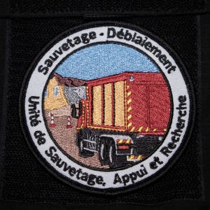 Può includere: Toppa ricamata circolare con un camion di soccorso rosso e giallo, un edificio e il testo "Sauvetage - Déblaiement, Unité de Sauvetage, Appui et Recherche". Sfondo nero.