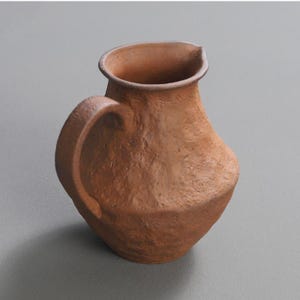 May include: A brown ceramic pitcher with a textured surface. The pitcher features a curved handle and a pouring spout. The earthenware vessel is likely used for holding liquids and has a classic design. The pitcher is made of earthenware.