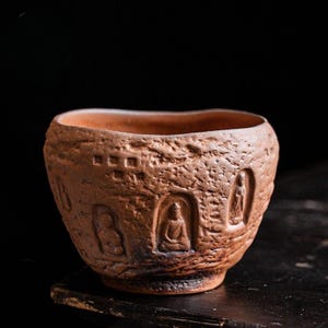 May include: A brown ceramic bowl with a textured exterior. The bowl features embossed images of seated figures within arched niches. The rim has a slightly irregular shape. The bowl is set against a dark background.