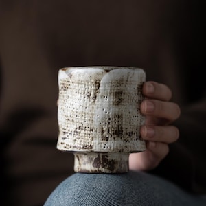 Puede incluir: Taza de cerámica hecha a mano con un esmalte blanco roto texturizado y motas marrones. La taza tiene una forma irregular única y se asienta sobre una pequeña base de pedestal. La taza es sostenida por una persona.