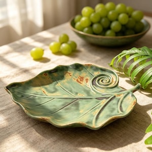 May include: A green ceramic leaf-shaped serving dish with a spiral design. The dish has a textured surface with brown accents and dark green veins. Grapes in a green bowl are in the background.