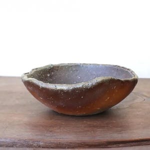 Puede incluir: Un pequeño cuenco de cerámica hecho a mano con un borde áspero e irregular. El cuenco tiene un exterior marrón oscuro con un borde más claro y texturizado. El interior es de un tono marrón más oscuro. El cuenco está sobre una superficie de madera.