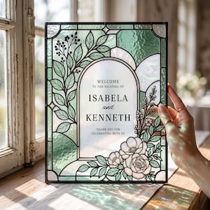 Op de afbeelding: Een rechthoekig trouwbord van glas-in-lood met een zwart metalen frame. Het midden bevat de tekst "WELCOME TO THE WEDDING OF ISABELA and KENNETH THANK YOU FOR CELEBRATING WITH US" omringd door groene glazen bladeren en witte bloemen.