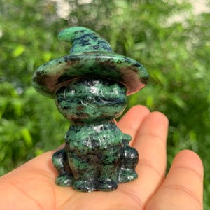 May include: A small carved stone figurine of a cat wearing a pointed hat. The sculpture is crafted from a mottled green and black mineral with dark inclusions, featuring a smooth, polished surface and a distinct feline shape with a wide brimmed hat.