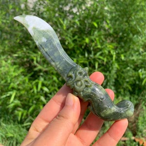 Könnte beinhalten: Ein geschnitzter Dolch aus grünem Stein mit einer geschwungenen Klinge mit weißer Spitze und einem Griff, der kunstvoll in Form eines Fabelwesens mit markanten Gesichtszügen und einem eingerollten Ende gestaltet ist, mit natürlichen Mustern.