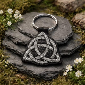 May include: A black leather keychain with a silver Celtic knot design. The keychain has a silver ring and is displayed on a dark gray stone surface with white flowers and greenery.