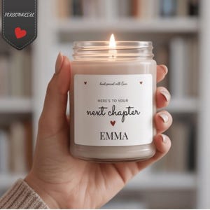 Può includere: Una candela accesa in un barattolo di vetro trasparente con un'etichetta bianca. L'etichetta riporta il testo "hand poured with love", "HERE'S TO YOUR next chapter" e il nome "EMMA" in nero, decorato con piccoli cuori rossi attorno al testo.